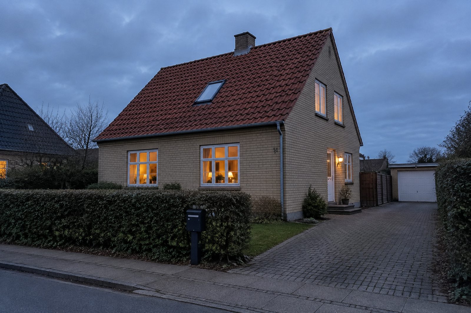 Dansk parcelhus med rødt tegltag og tændte vinduer i skumringen